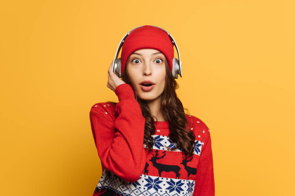 surprised girl in hat, wireless headphones and red ornamental sweater looking at camera isolated on yellow