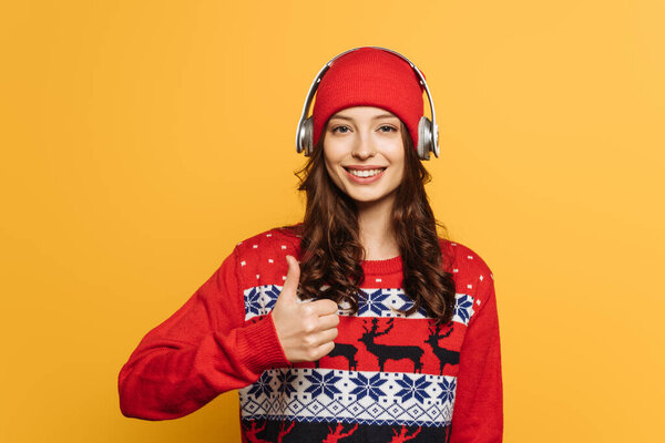 happy girl in wireless headphones on hat, in red ornamental sweater, showing thumb up isolated on yellow