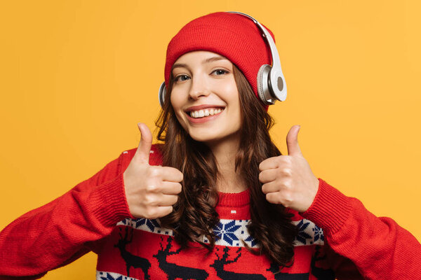 happy girl in wireless headphones on hat, in red ornamental sweater, showing thumbs up on yellow background