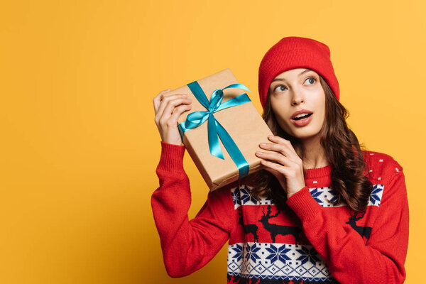 curious girl in hat and red ornamental sweater holding gift box on yellow background