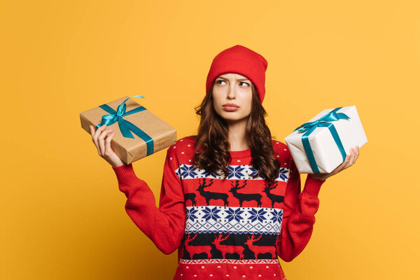 thoughtful girl in hat and red ornamental sweater holding gift boxes on yellow background