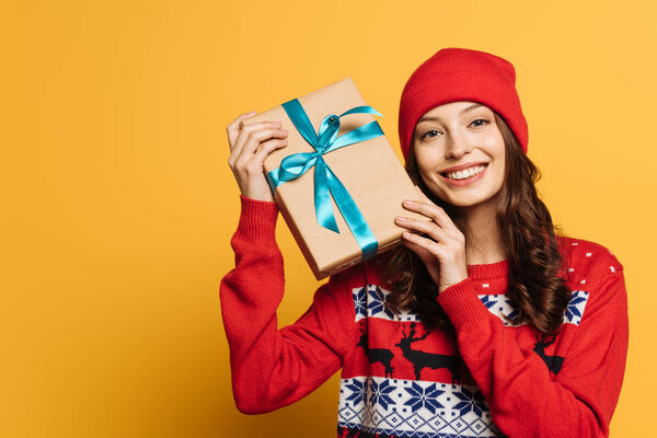 cheerful girl in hat and red ornamental sweater holding gift box on yellow background