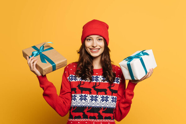 happy girl in hat and red ornamental sweater holding gift boxes on yellow background