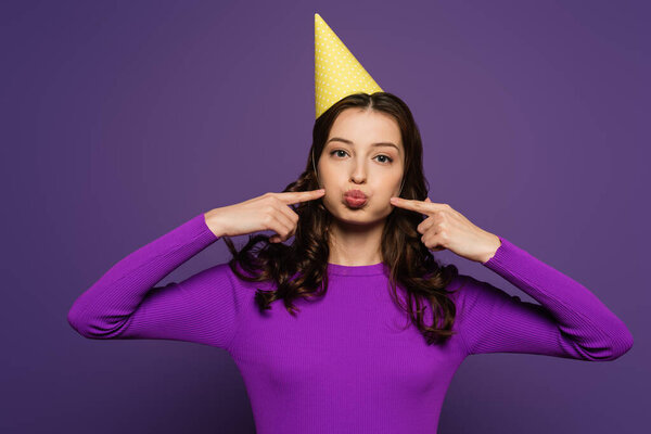 cheerful girl in party cap touching chicks with fingers on purple background