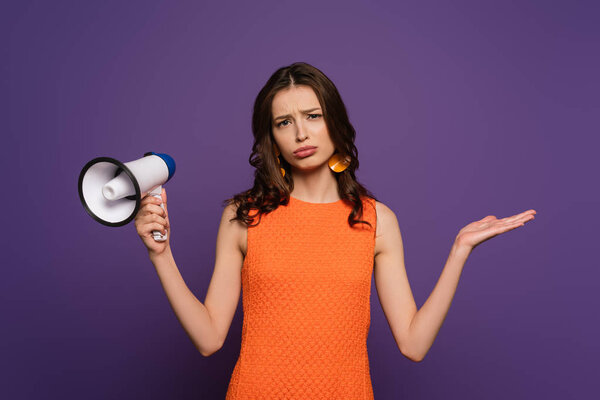 upset girl showing shrug gesture while holding megaphone and looking at camera isolated on purple