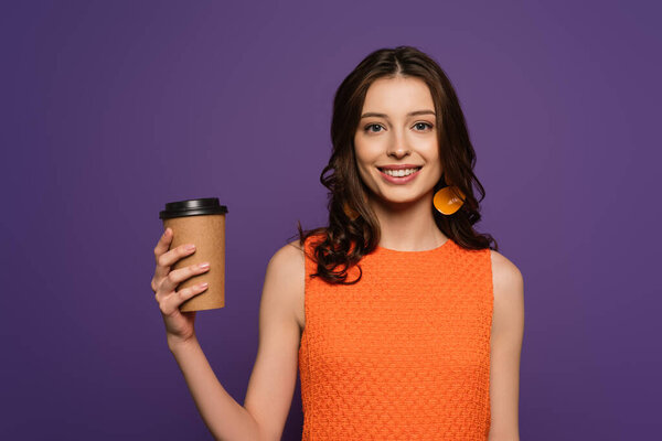 happy girl holding coffee to go and smiling at camera isolated on purple