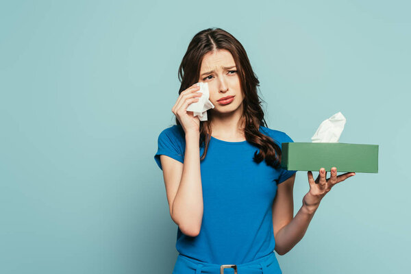 sad girl wiping tears with paper napkin while crying isolated on blue