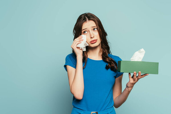 offended girl wiping tears with paper napkin while crying isolated on blue