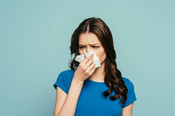 upset, diseased girl wiping nose with paper napkin isolated on blue