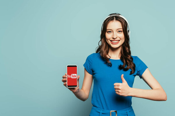 KYIV, UKRAINE - NOVEMBER 29, 2019: happy girl in wireless headphones showing thumb up while holding smartphone  with Youtube app on screen on blue background
