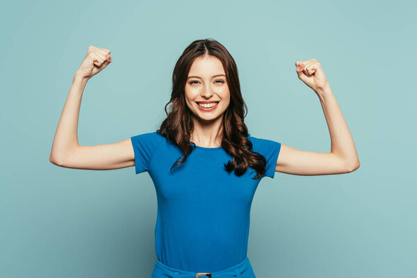 cheerful girl showing winner gesture while smiling at camera on blue background