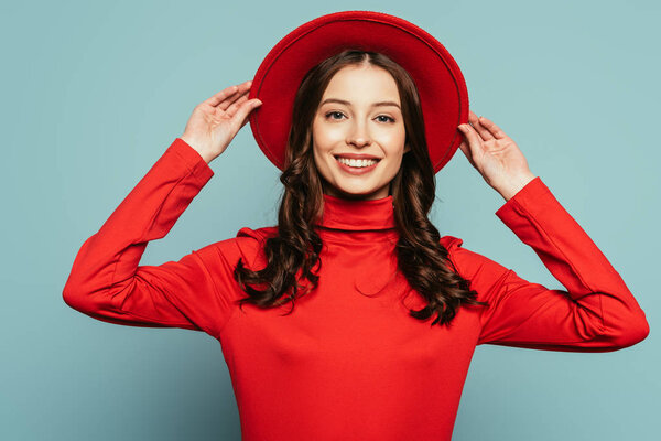 smiling stylish girl touching brims of hat while looking at camera on blue background