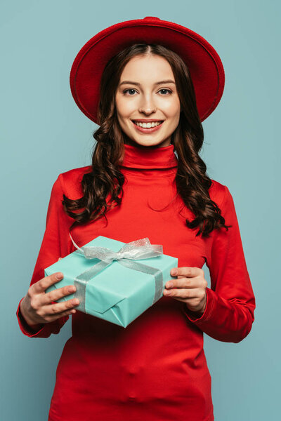 cheerful stylish girl smiling at camera while holding gift box isolated on blue