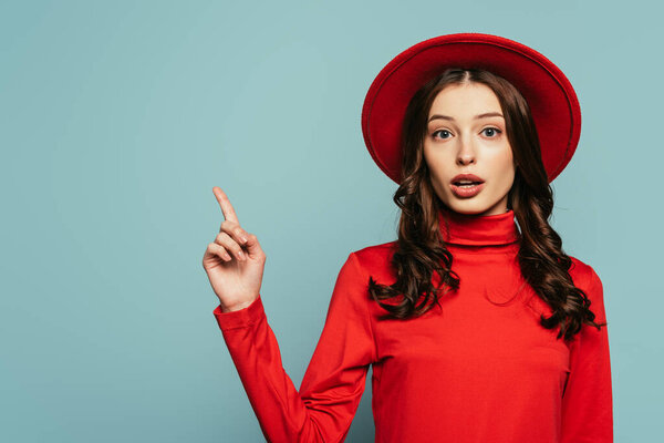 surprised stylish girl smiling at camera while pointing with finger isolated on blue