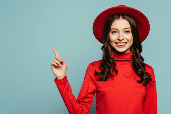 happy stylish girl smiling at camera while pointing with finger isolated on blue