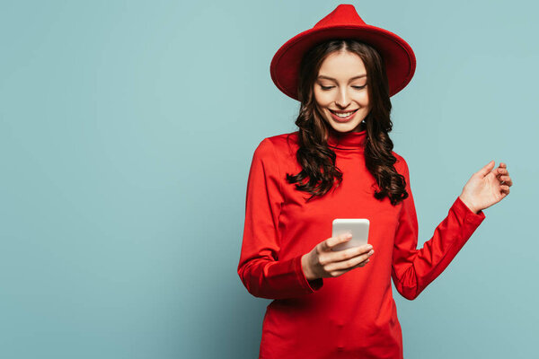 cheerful stylish girl smiling while chatting on smartphone on blue background