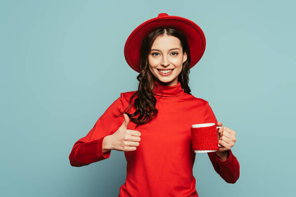 cheerful stylish girl showing thumb up while holding cup of coffee on blue background