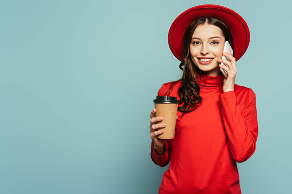 happy stylish girl talking on smartphone while holding coffee to go on blue background