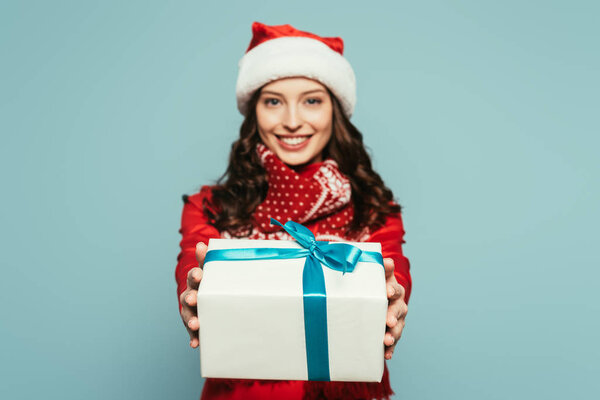 cheerful girl in santa hat showing gift box while looking at camera on blue background