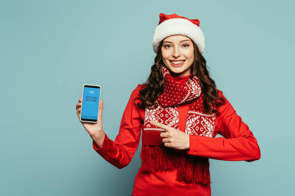 1KYIV, UKRAINE - NOVEMBER 29, 2019: happy girl in santa hat and red sweater pointing with finger at smartphone with Skype app on screen on blue background