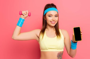 Gülümseyen sporcu kadın pembe arka planda dambılla çalışırken boş ekranla akıllı telefonu gösteriyor
