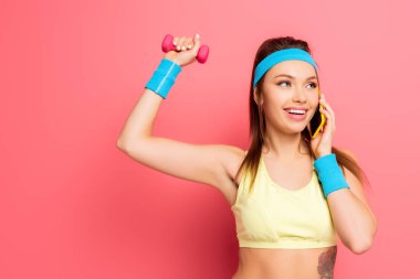 Gülümseyen sporcu kadın pembe arka planda dambılla çalışırken akıllı telefondan konuşuyor.