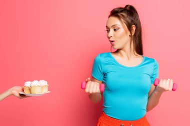 surprised sportswoman exercising with dumbbells near female hand with plate of delicious cupcakes on pink background clipart