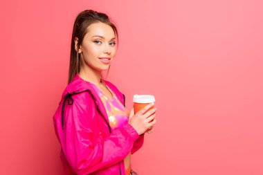 pretty, smiling sportswoman holding coffee to go while looking at camera on pink background clipart