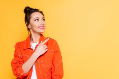 attractive smiling woman looking away and pointing with finger isolated on yellow clipart