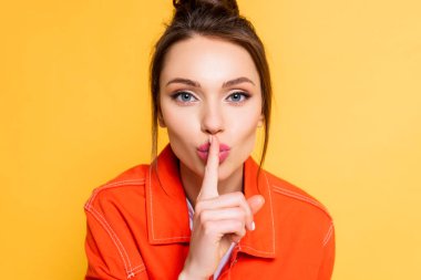 attractive young woman showing hush gesture while looking at camera isolated on yellow clipart