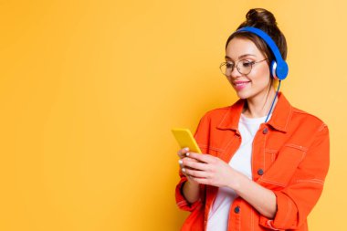 Kulaklıklı gülümseyen öğrenci sarı arka planda akıllı telefondan sohbet ediyor.