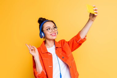 Kulaklıklı neşeli öğrenci akıllı telefondan selfie çekerken sarı renkte zafer işareti gösteriyor.