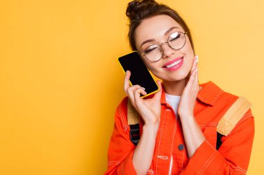 Gözlüklü gülümseyen öğrenci elinde akıllı telefonuyla boş ekranla sarı arka planda gözleri olan.
