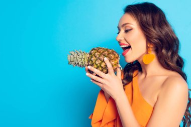 Neşeli kız, ağzı açık, mavi arka planda olgun ananas tutuyor.