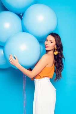Güzel stil sahibi kız kameraya gülümserken mavi arka planda büyük balonlar tutuyor.