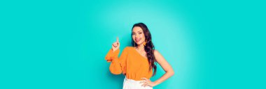 Neşeli, stil sahibi bir kızın el ele tutuşup turkuaz arka planda durduğu panoramik bir fotoğraf.