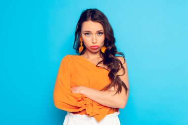 Gücenmiş, somurtan kız mavi arka planda çapraz kollarla kameraya bakıyor.