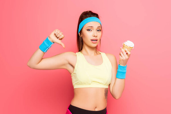 young sportswoman showing disapproval gesture while holding cupcake on pink background
