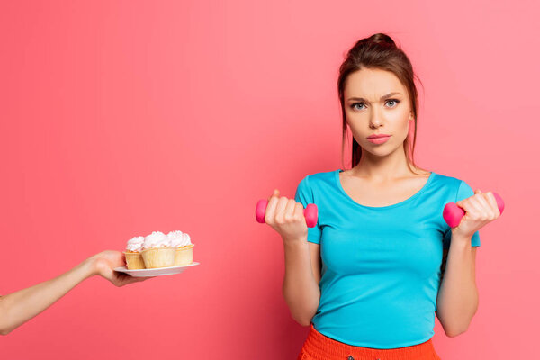 confident sportswoman exercising with dumbbells near female hand with plate of delicious cupcakes on pink background