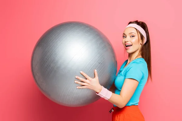 快乐的女运动员拿着银健身球 对着镜头笑着 背对着粉色的背景 — 图库照片