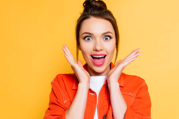 shocked young woman holding hands near face while looking at camera with open mouth isolated on yellow