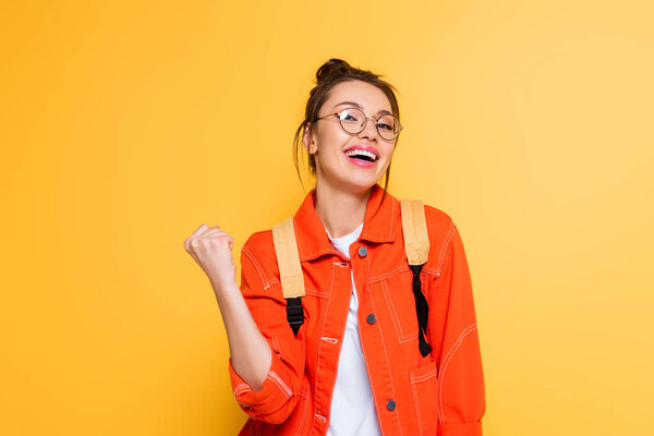 excited student in eyeglasses showing winner gesture while smiling at camera isolated on yellow