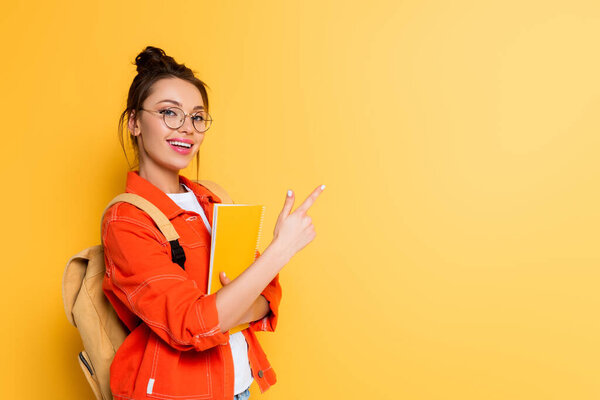 cheerful student holding notebook and pointing with finger on yellow background