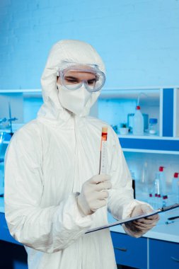 Tehlikeli madde giysisi ve gözlüklü bilim adamı elinde koronavirüs harfleri ve pano ile test tüpü tutuyor.