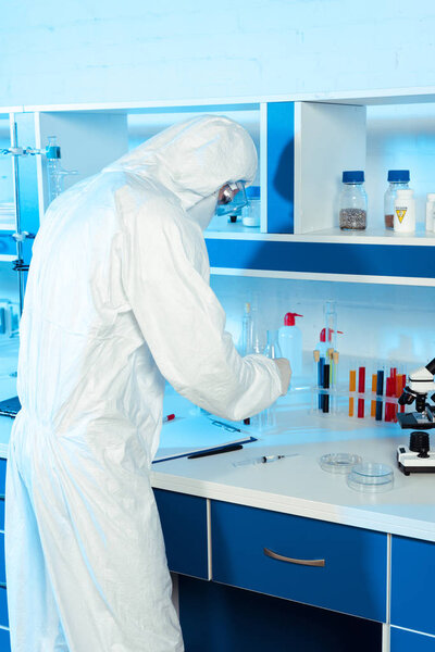 scientist in hazmat suit standing near syringe, clipboard and microscope 