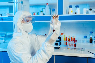 scientist in hazmat suit and latex gloves holding test tube with coronavirus lettering  clipart