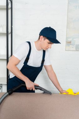 young man in cap and overalls removing dust on sofa with vacuum cleaner and cleaning cloth clipart