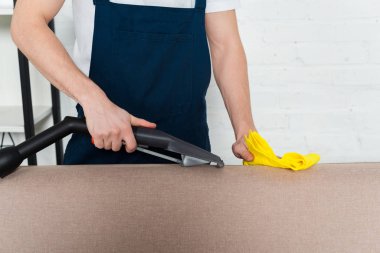 Elektrikli süpürge ve temizlik beziyle kanepeyi temizleyen adam manzarası.