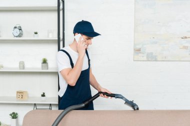 Şapkalı ve tulumlu temizleyici elektrikli süpürgeyle kanepeyi temizliyor ve akıllı telefondan konuşuyor. 