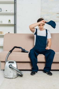 exhausted cleaner in overalls holding cap while sitting on sofa near vacuum cleaner  clipart